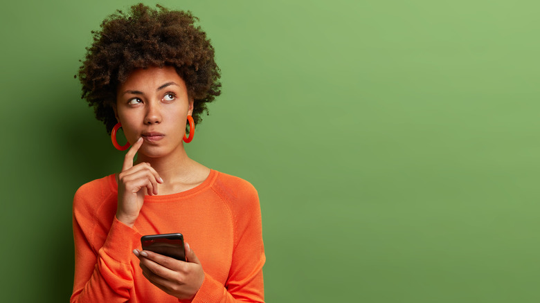 Woman wearing orange looking up thinking while holding her smartphone