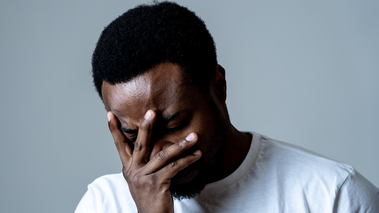 A man in a white shirt against a gray background holding his face 